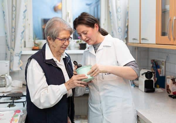 Mitarbeiterin des Besuchsdienstes unterstützt eine ältere Frau in der Küche bei einer alltäglichen Tätigkeit.